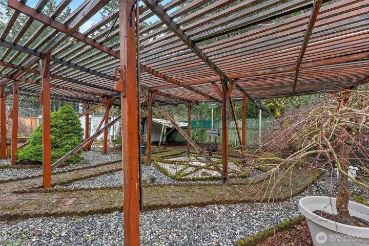 An expansive wood pergola anchors the backyard, framing a low-maintenance gravel garden with mature evergreens, decorative moss-edged pathways, and a storage shed tucked in the corner.
