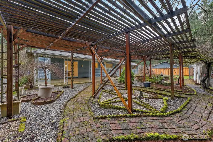 A wide view of the sprawling wood pergola showcases the scale of this outdoor space, with moss-edged brick pathways, gravel beds, and a direct sightline back to the covered patio and home.