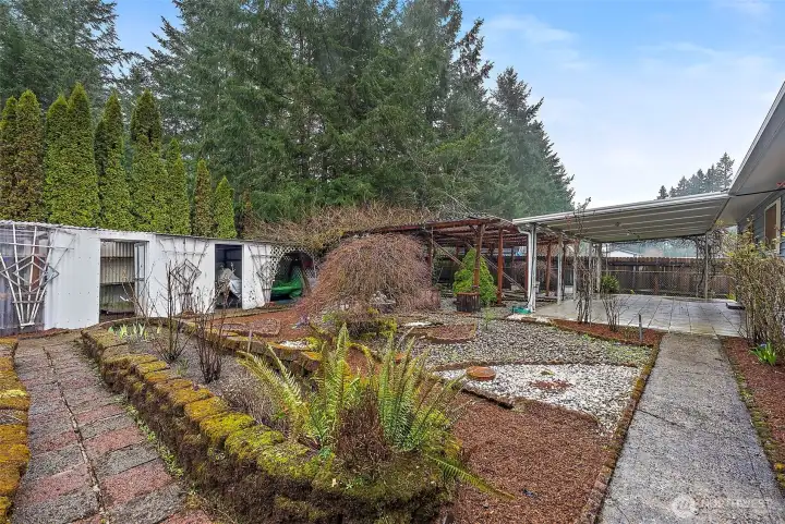 Raised stone-edged planting beds, established evergreens, and a gravel garden area surround the covered patio and greenhouse, offering a private and mature outdoor retreat.