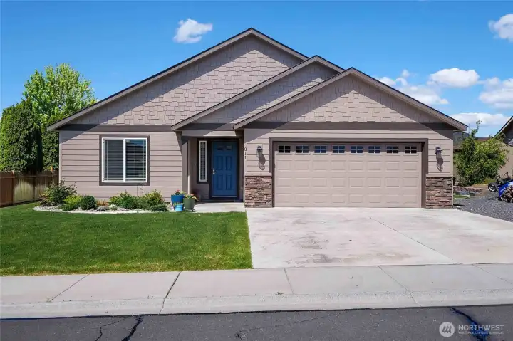 Like new, single-level 1,783 sq ft, 3-bedroom, Alderbrook home with gabled roof, stone accents, blue door, two-car garage, and neatly landscaped yard.