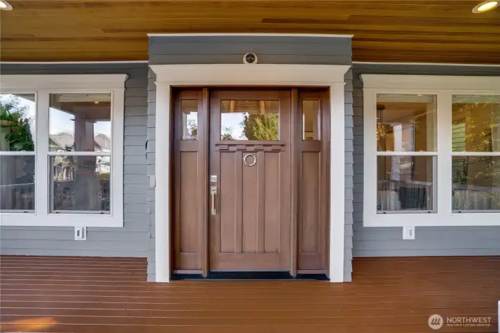 A solid wood craftsman door with glass insets anchors the wraparound porch. Remote-controlled blinds are visible through the flanking windows. Every detail at the entry was considered — from the hardware to the porch ceiling above.