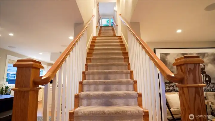 The grand staircase — oak newel posts, carpet runner, and a sight line rising three full floors — anchors the entry and immediately communicates the scale of this home. To the left, the dining room and kitchen. To the right, the living room, main floor bath, and Murphy bed office.