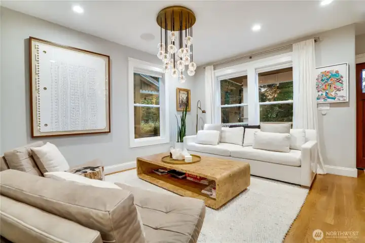 Light-filled living room with wide-plank oak hardwood floors, an oversized statement chandelier, and large-format fine art. In-ceiling surround sound speakers are integrated throughout. The open layout connects naturally to the dining room and kitchen beyond.