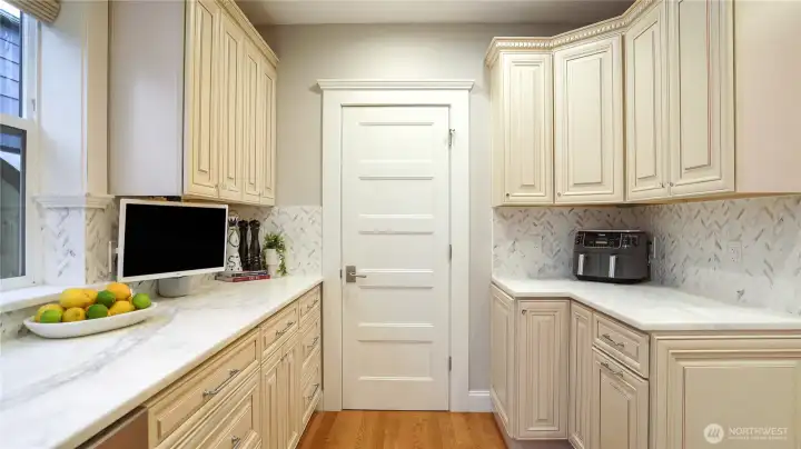 A full butler’s pantry with herringbone marble backsplash, Calacatta counters, upper and lower custom cabinetry on both sides, built-in TV, and a beverage refrigerator. This is a working prep kitchen — not a closet with shelves. It keeps the main kitchen clear when entertaining at scale.