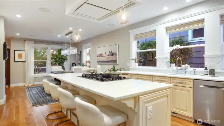 The open connection between kitchen and dining is the heart of the main floor. Island seating, the crystal linear chandelier beyond, and the porch visible through the dining room windows create a seamless flow from cooking to gathering. Wide-plank oak hardwood runs continuously throughout.