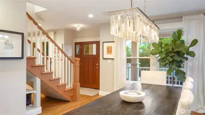 The dining room connects directly to the entry foyer, with the walnut craftsman front door and staircase visible beyond. The crystal linear chandelier anchors the space. Floor-to-ceiling sheer curtains frame the balcony windows. This room seats twelve comfortably and flows naturally to the kitchen on one side and living room on the other.