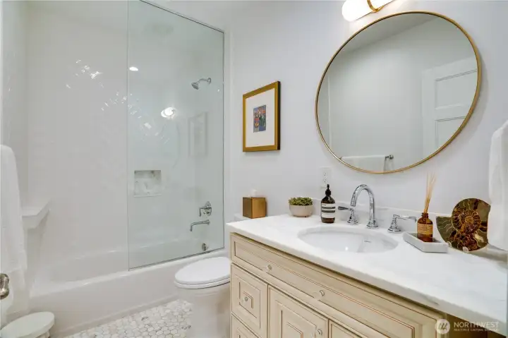 The main floor full bath features Carrara marble counters, a custom glazed vanity with gold-framed round mirror, herringbone tile shower surround, and hex tile floors. Styled with the same attention to finish detail as every other bath in this home. Serves both the main floor bedroom/office and guests.