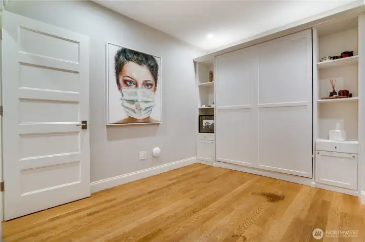 The main floor Murphy bed and office flex space — built-in cabinetry, flanking shelving, wide-plank oak floors, and a standing desk setup. Closed, it reads as a private office. Open, it’s a guest suite. For the executive or medical professional who needs both, this room delivers without requiring a choice.