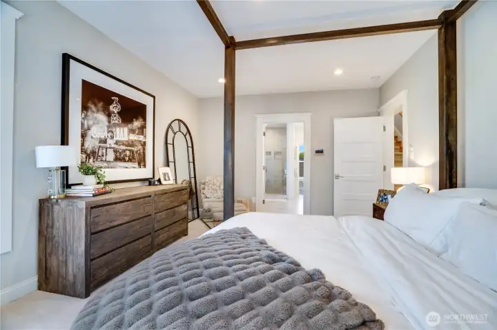 The sight line from the primary bedroom through to the en suite bath confirms what the rest of the home promises — every transition is intentional. The four-poster bed, arched mirror, and chaise create a layered suite experience. The door to the right leads to dual walk-in closets.