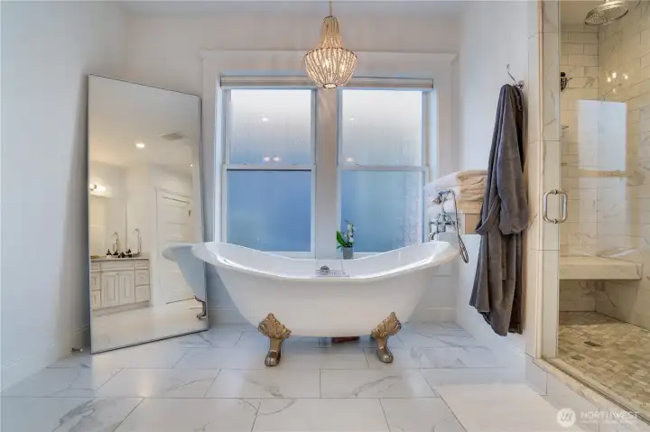 A freestanding clawfoot soaking tub with antique brass claw feet, beaded chandelier, frosted privacy windows, and Calacatta marble floors. The walk-in shower with Rohl fixtures is visible right. This is the photo that will stop buyers mid-scroll. Nothing in this price range delivers this moment.