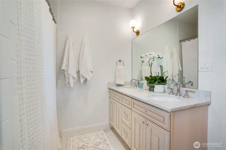 The second floor full bath — Carrara marble counter, double sink vanity with custom glazed cabinetry matching the primary, Rohl faucets, brass sconce, and orchid styling. The same finish standard carried from the primary suite down to every shared bath in this home.