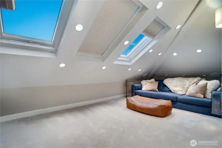 The third floor media room with three skylights, vaulted ceiling, recessed lighting throughout, and a blue leather sectional. On a clear day, the skylights pull in open sky. On a movie night, the blackout blinds close and the surround sound takes over. This room was designed for both.