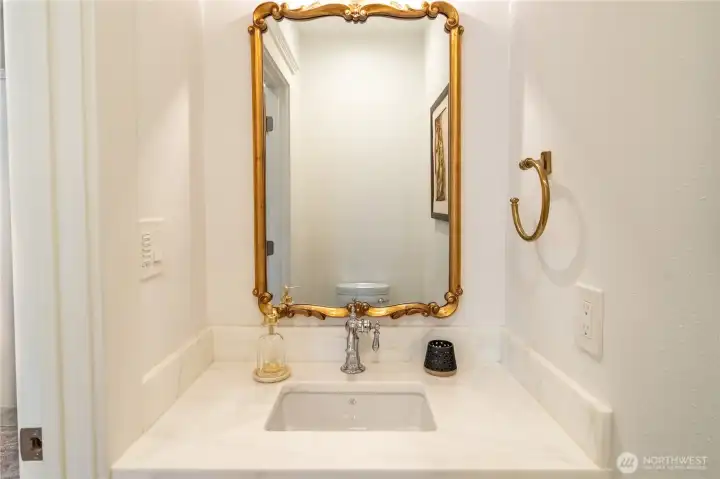 The third floor 3/4 bath vanity — an ornate gold-framed mirror, Rohl bridge faucet, Carrara marble counter, brass towel ring, and matching custom glazed cabinetry. The same finish standard carried from the primary suite all the way to the top floor. In a home with this level of detail, even the third floor bath makes a statement.