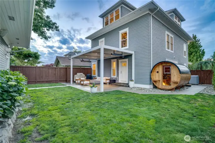 The rear elevation at dusk — warm light in every window, StruXure pergola left, cedar barrel sauna glowing right, green lawn, and three floors of architectural presence. This photo captures what it feels like to come home. The outdoor living sequence begins here.