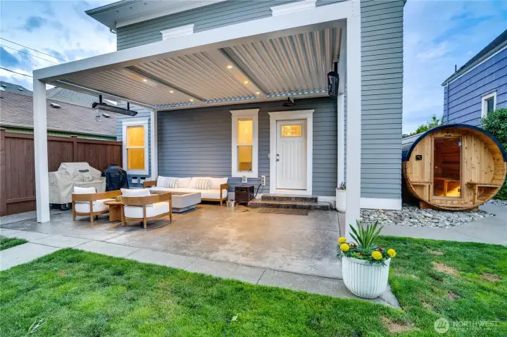 The StruXure louvered pergola with integrated recessed lighting, infrared heaters, teak outdoor furniture, and a concrete gas fire pit. The louvered roof opens and closes on demand — sun when you want it, cover when you don’t. The cedar barrel sauna is visible right, completing the outdoor circuit.