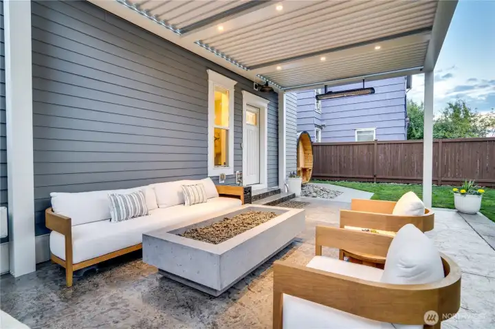 Teak chairs, a concrete linear fire pit, and the louvered StruXure ceiling above — this is the outdoor living room. Infrared heaters extend the season. The sealed concrete floors, fenced yard, and mature plantings create a private setting that feels removed from the neighborhood around it.