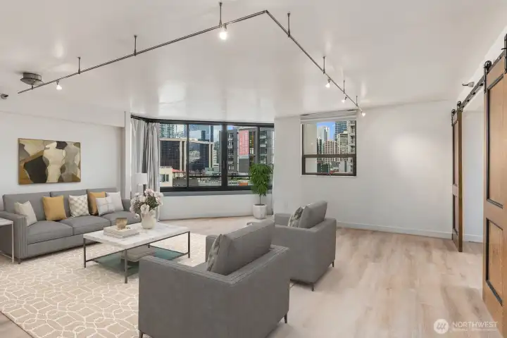 Virtually staged Seattle condo living area showcasing an open layout, modern track lighting, and large windows overlooking surrounding downtown buildings.