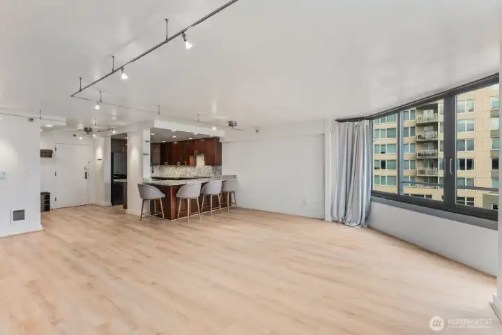 Bright urban condo living space with expansive windows overlooking surrounding city buildings and an open kitchen with peninsula seating in the background.