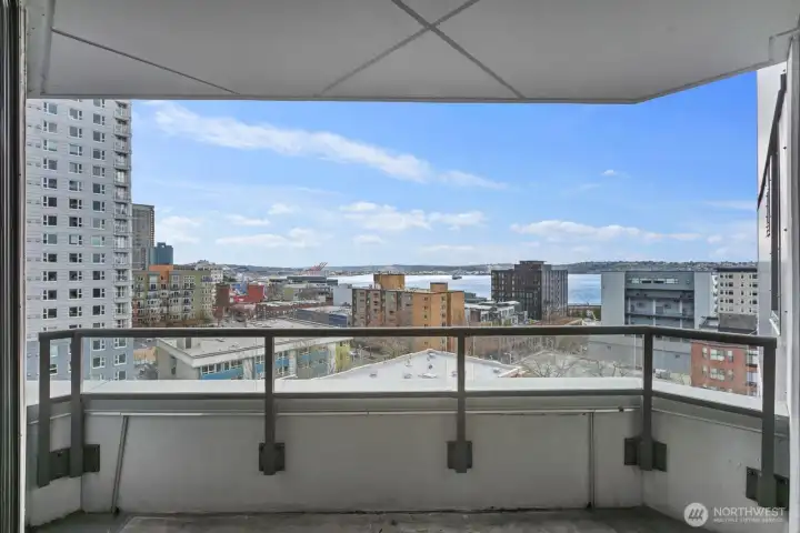 Private Seattle condo balcony overlooking surrounding city buildings with expansive Puget Sound water views and open sky beyond the urban skyline.