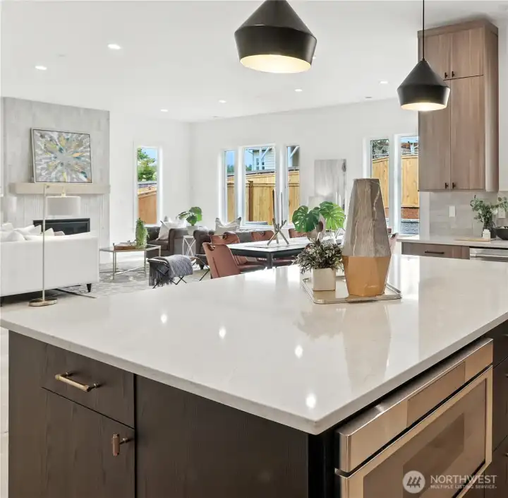 A striking centerpiece for daily life and entertaining alike. The chef’s kitchen pairs rich wood cabinetry with sleek black-and-brass accents, grounded by an oversized quartz island that doubles as a prep station and social hub. Pendant lighting adds modern warmth, while full-height windows draw natural light across the open-concept layout. The seamless flow into the great room highlights the home’s sense of space and connection — perfect for casual gatherings or elegant evenings by the fire.