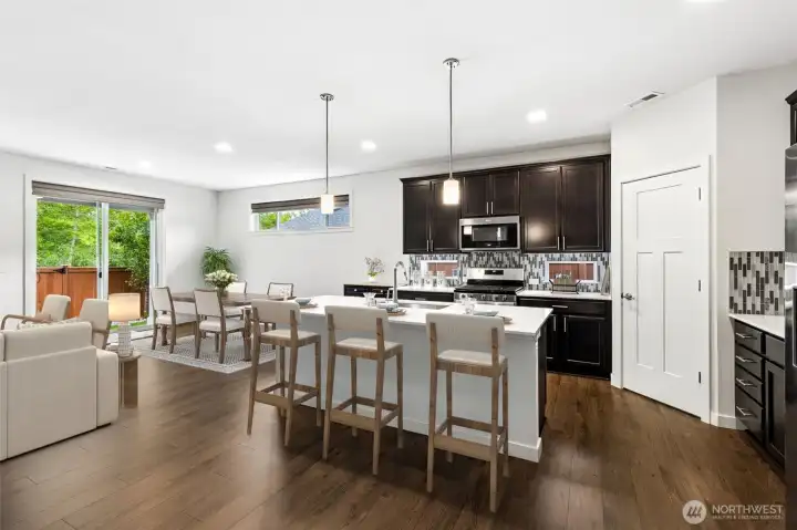 The heart of the home is this beautiful open-concept great room where the kitchen, dining area and living spaces all flow together effortlessly, making it easy to stay connected whether you're hosting friends or simply enjoying a quiet evening at home. The oversized island offers comfortable bar seating and quickly becomes the place where everyone naturally gathers, while handsome dark shaker cabinetry creates rich contrast against the warm flooring below. Two stylish pendant lights anchor the island and recessed lighting throughout the space keeps everything bright and welcoming. From this angle you can also spot the generous walk-in pantry tucked conveniently nearby, making everyday life easier.
