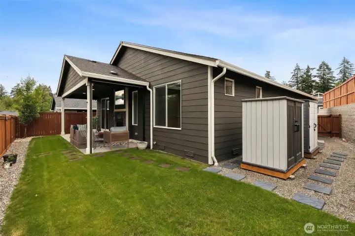 This backyard strikes a wonderful balance between low-maintenance living and usable outdoor space. You'll notice the two included storage sheds tucked neatly along the side of the home, connected by thoughtful paver pathways that help keep your feet out of wet grass during our Pacific Northwest seasons. The generous level lawn offers plenty of flexibility for whatever your outdoor dreams may be, whether that's a putting green, raised garden beds, a peaceful water feature or even a future hot tub installation. One particularly smart upgrade is the remote-controlled solar shade installed along one side of the covered patio, helping block wind, rain and afternoon sun while also trapping warmth from the outdoor fireplace for a more comfortable year-round outdoor living experience. It's the kind of backyard that's ready to adapt to whatever lifestyle you bring with you.