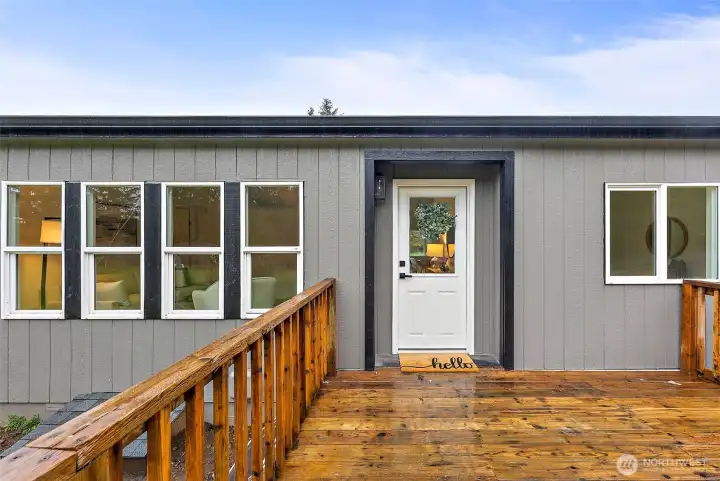 The front deck showcases a charming white entry door with glass panel and a "hello" doormat, framed by fresh board-and-batten siding with black accents.