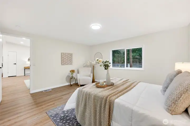 Another angle of the primary suite highlights the open, airy feel with LVP flooring, a bright window, and a private hallway leading to living areas.