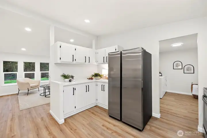Kitchen peninsula with white cabinetry and stainless refrigerator, open to the living room with views to the yard and a doorway leading to the utility/laundry room.