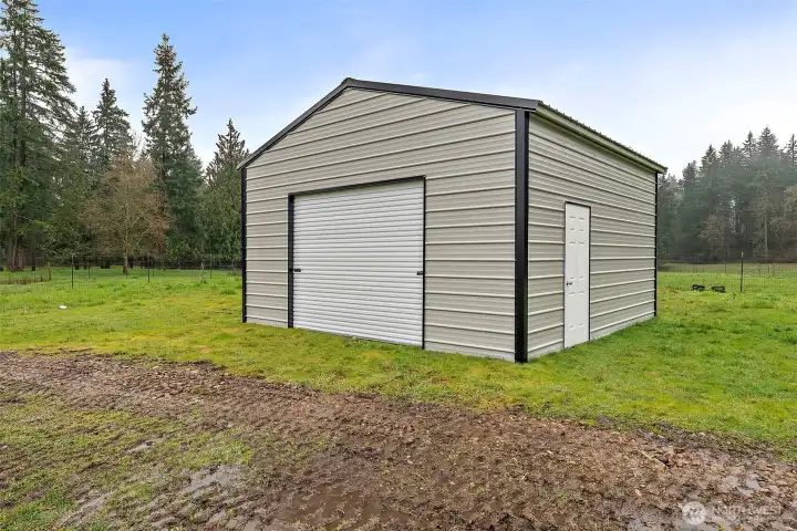 New spacious detached metal garage with a roll-up door and walk-in entry, surrounded by open yard and mature tree line — ideal for storage, a workshop, or vehicles.
