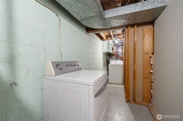 The lower-level utility room includes a washer and dryer with additional space for storage or future upgrades. Exposed ductwork and concrete walls highlight its functional, unfinished character.