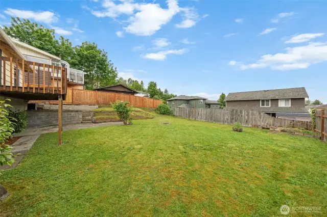 A wide view of the backyard highlights the elevated deck and fire pit, creating a perfect setup for entertaining or relaxing outdoors. The gentle slope and retaining wall add dimension and visual interest to the space.