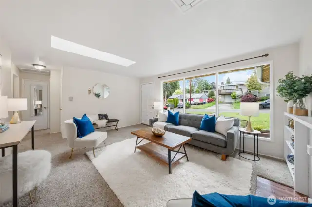 The large front window offers a full view of the landscaped front yard and quiet street beyond. Natural light enhances the open feel of the living room while keeping the outdoors in sight. To the left, a hallway leads to two bedrooms and the main bathroom.