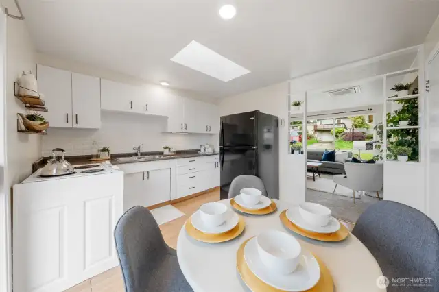 The eating space, kitchen, and living room are seamlessly connected, creating an open layout ideal for entertaining. A skylight and pass-through shelving enhance the natural light and sense of flow throughout the space.