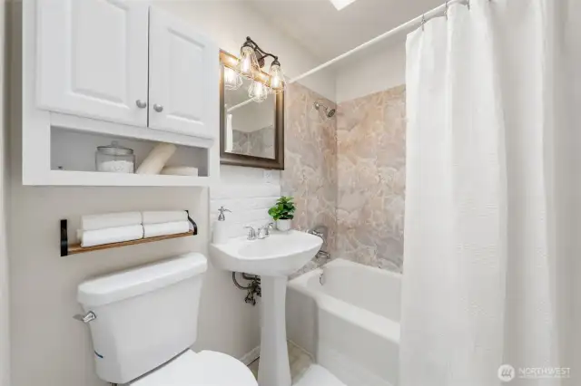 The main hall bathroom includes a pedestal sink, built-in storage, and a full tub with a marble-look surround. A skylight above the shower adds natural light to the clean, functional design.