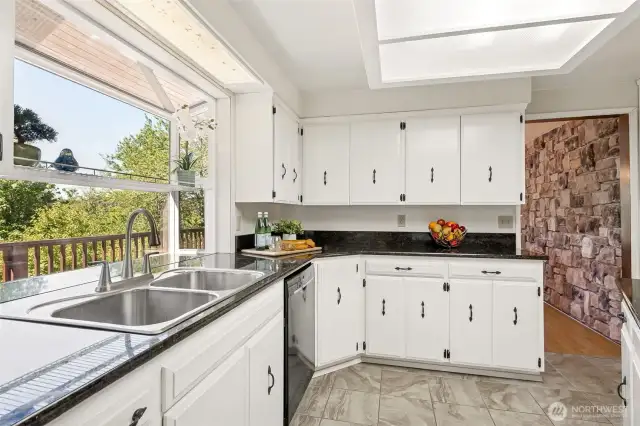The garden window over the sink isn’t just for herbs, it overlooks 110 acres of protected Capitol Land Trust forest and your own wraparound deck. It’s the kind of view that turns dishwashing into a design moment. Dark countertops ground the space, and through the doorway, the stone fireplace wall wraps back in, quietly tying the entire layout together.