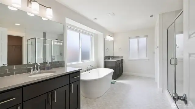 Step into this spacious en suite bathroom featuring separate vanities, a soaking tub, a walk-in shower, large mirrors, and plenty of cabinet space