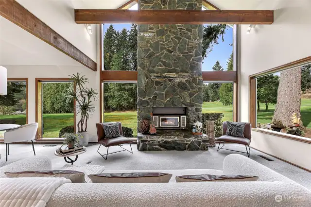 The second living room area with stone fireplace surrounded by large windows with serene views and wildlife.