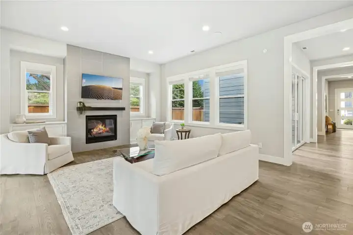 The open-concept living area is bathed in natural light from oversized windows, highlighting the home’s airy proportions and refined finishes. A contemporary gas fireplace with sleek tile surround creates a warm focal point, perfect for gathering or relaxed evenings at home.