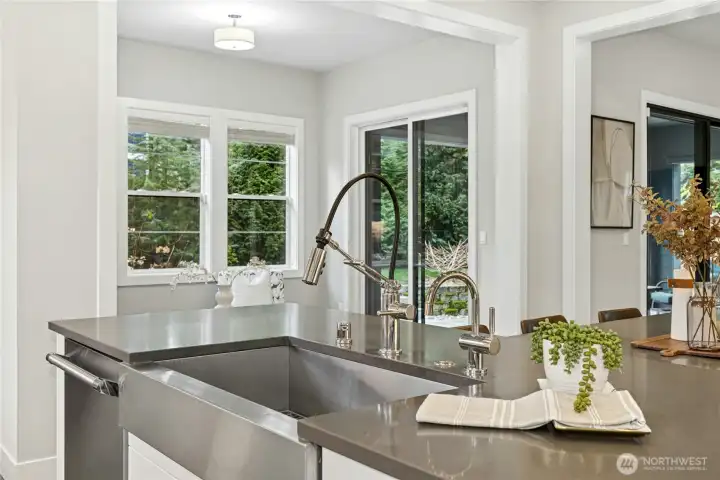 The expansive island features sleek quartz countertops and a statement stainless steel farmhouse sink with professional-grade fixtures. Positioned to overlook the nook and patio beyond, this workspace combines style and function while keeping you connected to both indoor and outdoor living spaces.
