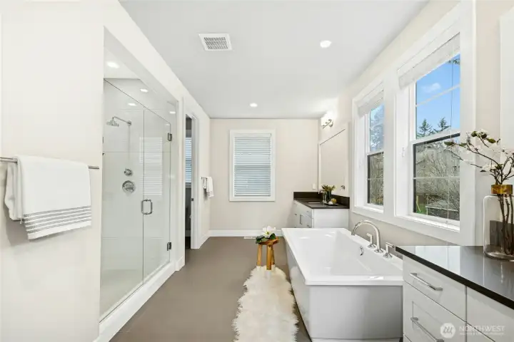 The spa-inspired primary bath offers a serene escape, featuring a soaking tub framed by expansive windows that invite natural light. A glass-enclosed shower with modern tile surround and dual vanities with quartz countertops create a refined and functional retreat designed for everyday luxury.