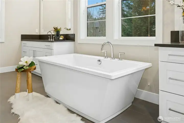 A closer view of the freestanding soaking tub highlights the spa-like atmosphere of the primary bath. Positioned beneath large windows, the tub is complemented by modern fixtures and nearby dual vanities, creating a tranquil space designed for relaxation and everyday comfort.