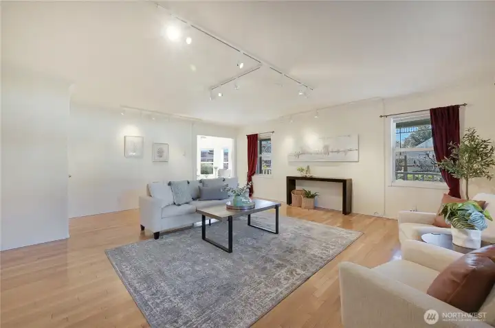 Light-filled living room with hardwoods, updated track lighting, and wall space for art—three picture windows overlook mature landscaping and fenced yard.