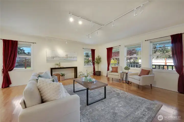 Bright, light-filled living room with hardwood floors, updated track lighting, and space to showcase art—three picture windows look out to mature landscaping and the fenced yard.