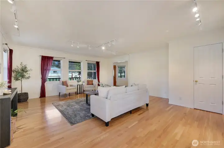 Light-filled living room with hardwoods, updated track lighting, and wall space for art—three picture windows overlook mature landscaping and fenced yard.
