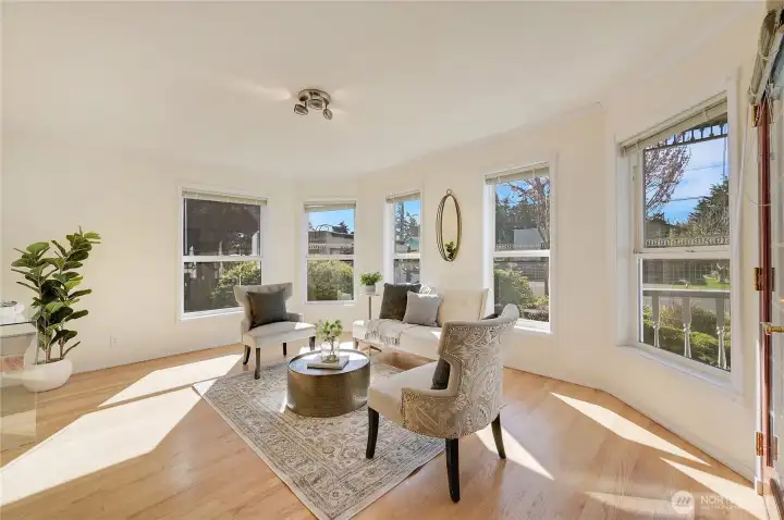 Bright bayed sitting room with wraparound windows, warm wood floors, and peaceful front-garden views—ideal for morning coffee, reading, or intimate conversations.