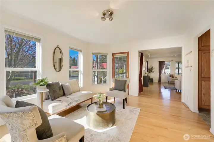 Light-filled bayed sitting room with wraparound windows and warm wood floors, overlooking the front garden—an inviting spot for morning coffee, reading, or conversation.