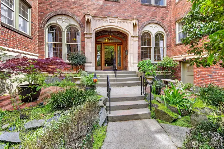 A storybook entrance awaits — lush gardens, ornate stonework, and a grand arched entry set the tone for life at Queen's Court, Seattle's beloved 1930 prewar landmark in Lower Queen Anne.