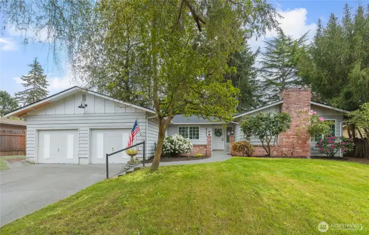 Classic ranch-style home with board-and-batten siding, brick chimney, and mature evergreen backdrop. Lush lawn and blooming rhododendrons add excellent curb appeal.