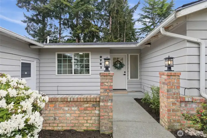Welcoming brick pillar entry with lantern-style lighting, wreath-adorned front door, and a charming paved walkway framed by flowering landscaping.